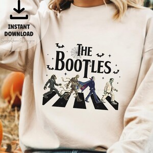 May include: Beige sweatshirt with a Halloween-themed graphic. The design features the text "The Bootles" with illustrations of classic monsters crossing a crosswalk. Includes bats, a spiderweb, and stars.