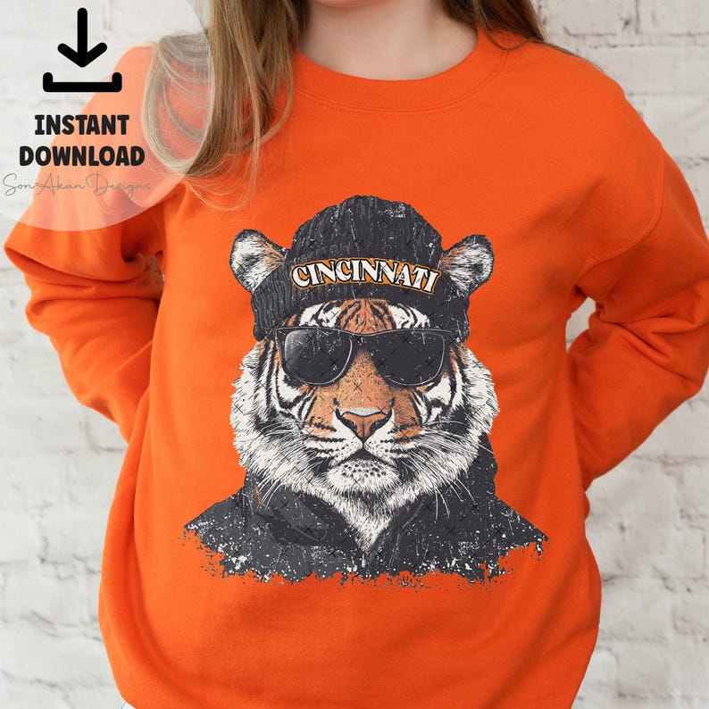 May include: Orange sweatshirt featuring a graphic of a tiger wearing a black beanie with the word "CINCINNATI" on it and sunglasses.