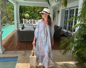 May include: A woman wearing a light blue and white patterned kaftan dress with a straw hat walks by a swimming pool. She is carrying a white tote bag.