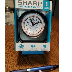 May include: Black analog alarm clock with a white face and red and white checkered design. The clock face has the text "Start Your Day With" and lists items like "Good Friends", "Hot Coffee", "and a Diner Breakfast". The clock is battery operated and has an easy to read dial, ascending alarm volume, and on demand backlight.