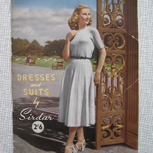 Puede incluir: Portada de revista vintage con una mujer con un vestido azul claro con falda plisada, mangas cortas y cinturón negro. El texto "Dresses and Suits by Sirdar" es visible. La mujer está cerca de una puerta ornamentada.