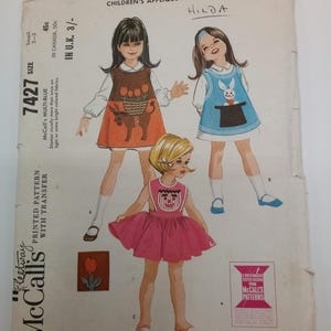 Könnte beinhalten: Vintage McCall's Nähmuster 7427 für Kinder-Schürzen mit Applikationen. Das Muster enthält drei verschiedene Designs: ein Kaninchen, einen Esel mit einem Blumenkorb und ein Clownsgesicht. Das Muster ist auf weißem Papier mit blauem und schwarzem Text gedruckt. Das Muster ist für die Größen 2-3 und kostet 45 Cent.