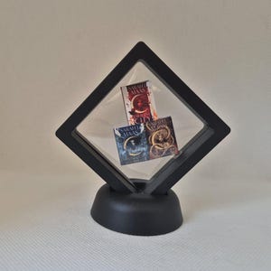 Op de afbeelding: Een zwarte display case met drie miniatuurboeken met kleurrijke covers. De boeken heten "Crescent City", "House of Earth and Blood" en "Throne of Glass". De case heeft een doorzichtige voorkant en een zwarte basis.