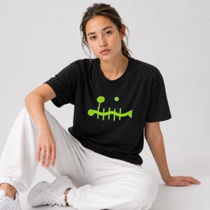May include: A black short-sleeved t-shirt featuring a lime green graphic of a stylized fish skeleton with a circular shape above it. The shirt has a crew neckline and is paired with loose-fitting white sweatpants and white athletic sneakers.