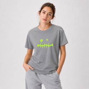 Peut inclure: Un t-shirt gris à manches courtes arborant un motif graphique vert citron vif représentant une arête de poisson stylisée sur la poitrine. Le design comprend une colonne vertébrale horizontale avec des côtes, une queue triangulaire et deux cercles.