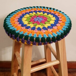 May include: A wooden stool with a colorful crocheted seat cushion. The cushion is made with a variety of bright colors, including orange, yellow, green, blue, and pink. The cushion has a circular design with a center circle and concentric rings of color.