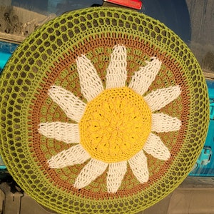 May include: A crocheted daisy flower design in shades of green, brown, white, and yellow. The design is circular and has a textured, woven appearance.