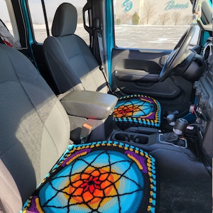 May include: A blue Jeep interior with two crocheted seat covers. The seat covers are colorful and feature a mandala design. The driver's seat cover is blue, orange, yellow, and purple. The passenger seat cover is blue, orange, and yellow.
