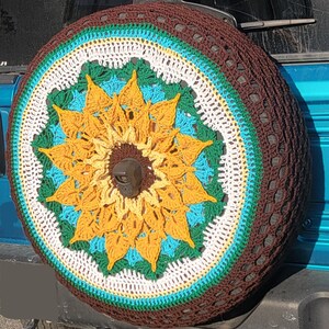 May include: A crocheted sunflower tire cover in brown, white, yellow, green, and blue. The cover is made of yarn and has a crocheted design.