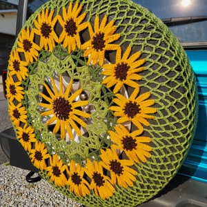 May include: A green crocheted tyre cover with a sunflower design. The cover is made of a soft, crocheted material and features a repeating pattern of sunflowers.