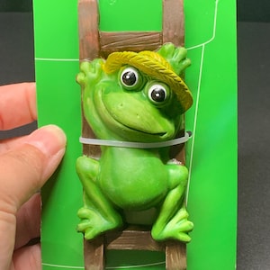 May include: A green frog figurine climbing a brown wooden ladder, wearing a yellow straw hat. The frog has large black eyes and is attached to a green backing card with a white zip tie. A whimsical garden decoration.