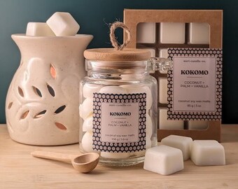 Puede incluir: Un tarro de vidrio lleno de pastillas de cera blancas, etiquetado como "Kokomo Coconut Palm Vanilla" por "auri candle co.". El tarro tiene una tapa de madera y está sentado sobre una superficie de madera con una cuchara de madera y otras pastillas de cera.
