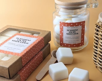 Puede incluir: Dos cajas de cera para fundir con la etiqueta "Sunshine on My Shoulder" y el aroma "Naranja, Coco, Flor de Algodón". Las cajas están apiladas una encima de la otra. Hay un frasco de vidrio lleno de cera para fundir y una cuchara de madera con cera para fundir sobre una superficie naranja claro.