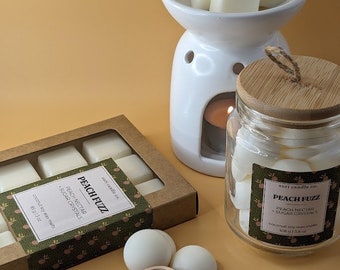 Puede incluir: Un juego de fundentes de cera en una caja marrón con una etiqueta verde y blanca que dice "Peach Fuzz Peach Nectar Sugar Crystals Coconut Soy Wax Melts 1.8 oz". La caja está al lado de un frasco de vidrio con una tapa de madera lleno de fundentes de cera blanca. El frasco tiene una etiqueta que dice "Peach Fuzz Peach Nectar Sugar Crystals Coconut Soy Wax Melts 1.08 oz". También hay un calentador de cera de cerámica blanca con una vela dentro y cuatro fundentes de cera blanca en la parte superior.