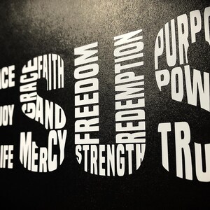 May include: Black background with white text spelling out the word "JESUS" in large letters.  The letters are made up of smaller words, including "love", "hope", "peace", "joy", "salvation", "life", "grace", "faith", "mercy", "and", "freedom", "redemption", "strength", "purpose", "power", and "truth".