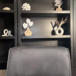 May include: A dark wood bookshelf filled with decorative items. The shelves display white coral, abstract sculptures, and wooden cutouts of plants in vases. A gray chair is in the foreground, and a diffuser sits on the bottom shelf.