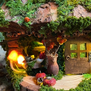Może przedstawiać: Miniaturowy domek dla wr&oacute;żek z dachem pokrytym mchem i drewnianymi drzwiami. Dom jest ozdobiony suszonymi kwiatami, grzybami i małymi czerwonymi jabłkami. Mała filiżanka i spodek stoją na stole. Scena jest oświetlona ciepłym, świecącym światłem.