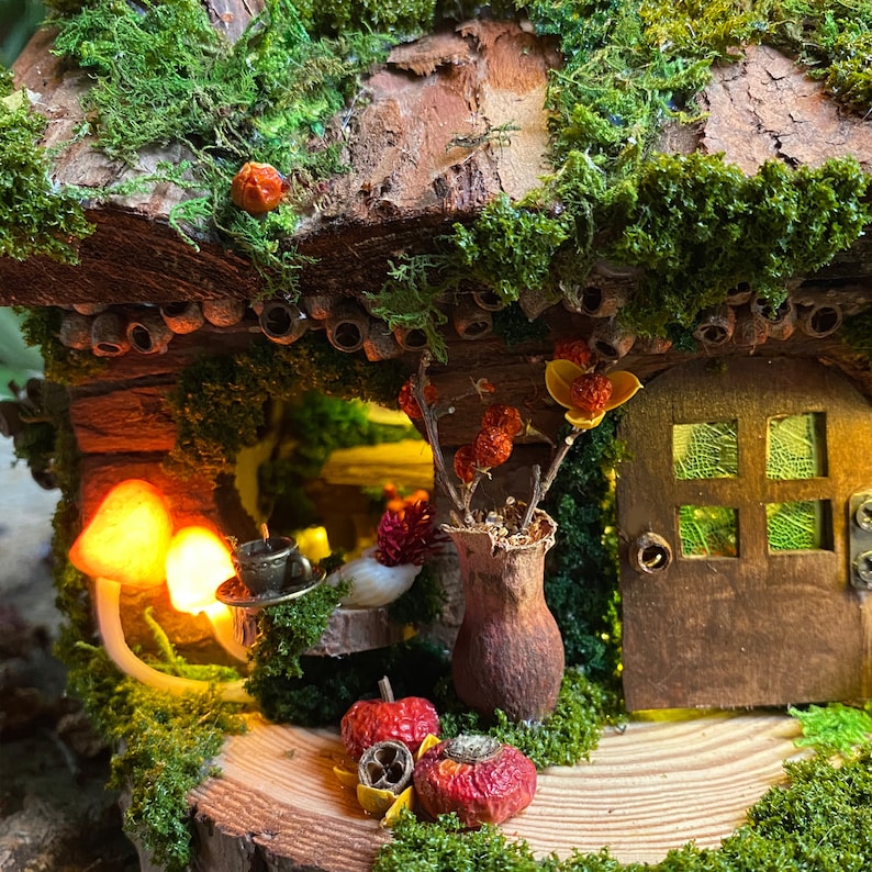 Może przedstawiać: Miniaturowy domek dla wr&oacute;żek z dachem pokrytym mchem i drewnianymi drzwiami. Dom jest ozdobiony suszonymi kwiatami, grzybami i małymi czerwonymi jabłkami. Mała filiżanka i spodek stoją na stole. Scena jest oświetlona ciepłym, świecącym światłem.