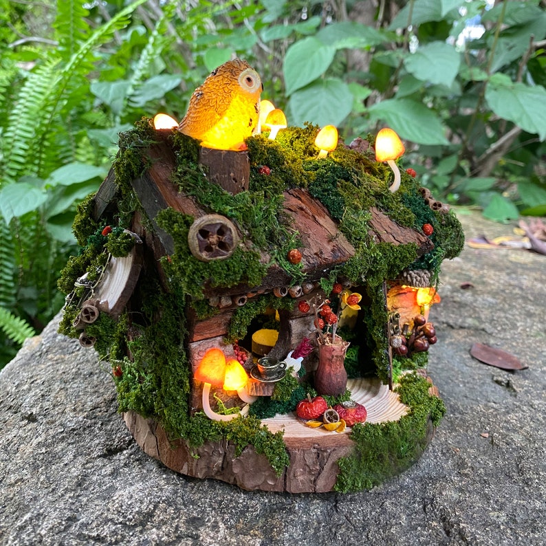 Może przedstawiać: Miniaturowy domek dla wr&oacute;żek z dachem i ścianami pokrytymi mchem, ozdobiony świecącymi grzybami i figurką sowy. Domek ma drewnianą podstawę i jest udekorowany małymi jagodami i naturalnymi elementami. Światła tworzą ciepłą, zachęcającą poświatę.