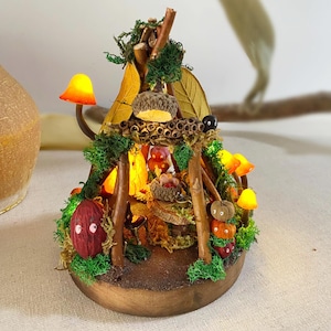 May include: A miniature woodland scene featuring a twig teepee, glowing lights, and autumnal decorations. The teepee is decorated with moss, leaves, and acorns. There are small pumpkins, mushrooms, and other woodland creatures inside the teepee.