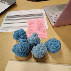 May include: Six blue crocheted balls on a wooden surface. The balls are soft and fluffy, and they are arranged in a cluster.