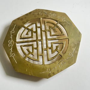 Puede incluir: Pieza decorativa octogonal de latón con un diseño geométrico central y delicados detalles florales. El latón tiene un tono dorado cálido y el diseño está recortado, revelando el fondo. Probablemente utilizado con fines decorativos.