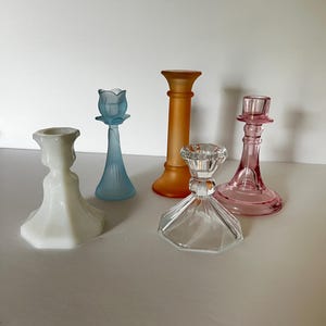 May include: A collection of five glass candle holders in various shapes and colours, including a white milk glass piece, a light blue tulip-shaped holder, a tall frosted orange pillar, a clear faceted glass base, and a pink glass candle holder.