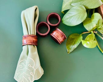May include: Three wooden napkin rings in a dark brown color. The rings are on a green surface with a green napkin with a white leaf design.