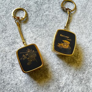 May include: Two vintage keyrings with gold-toned metal frames and key rings. One features a floral design on a black background. The other displays an Opel Coupe 1909 car illustration. Both have a gold chain link.