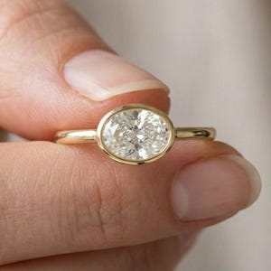 Könnte beinhalten: Ein goldener Ring mit einem großen, oval geformten Diamanten. Der Diamant ist in einer Zargenfassung gefasst, und das Band ist aus poliertem Gold. Der Ring wird von den Fingern einer Person gehalten.