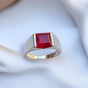May include: A silver ring featuring a large, square-cut, vibrant red gemstone. The ring has a simple, modern design with a polished finish, showcasing the gemstone's color and clarity. The ring is set against a white background.