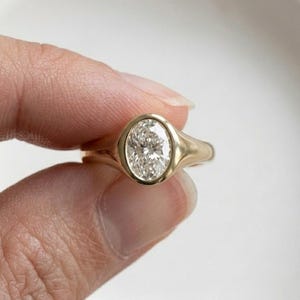 Op de afbeelding: Een gouden ring met een centrale, ovaalvormige, heldere edelsteen in een gladde zetting. De ringband is van massief goud met een gepolijste afwerking en loopt naar achteren toe iets taps toe. Het ontwerp benadrukt de ovale steen.