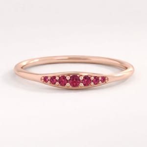 Puede incluir: Un anillo de oro rosa con una fila de siete pequeñas gemas redondas de color rojo brillante engastadas en una banda ligeramente curvada. El anillo se presenta sobre un fondo blanco liso, resaltando su diseño y color.