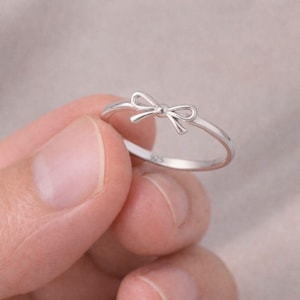 May include: A delicate silver ring featuring a bow design. The ring is crafted from shiny, polished metal and has a slender band. The bow is centered on the ring and adds a touch of elegance. The ring is held by a hand.