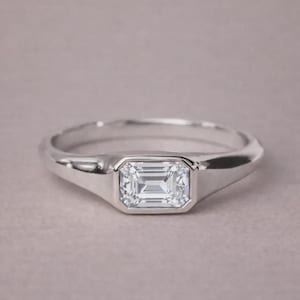 May include: A silver ring with a rectangular, emerald-cut gemstone. The ring has a smooth, polished band and a bezel setting that securely holds the clear, sparkling stone. The ring is displayed on a neutral background.