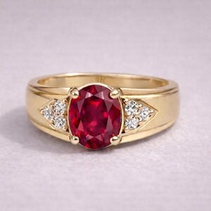 Puede incluir: Anillo de oro con un gran rubí ovalado en el centro. Pequeñas piedras claras están engastadas en un patrón triangular a cada lado del rubí. La banda es de oro pulido liso. Un diseño clásico.