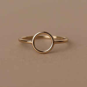 Könnte beinhalten: Ein minimalistischer Goldring mit kreisförmigem Design. Der Ring hat ein dünnes Band und einen markanten offenen Kreis in der Mitte. Der Ring besteht aus glänzendem, goldfarbenem Metall und befindet sich vor einem neutralen Hintergrund.