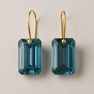 May include: Two teal-coloured, rectangular earrings with gold-coloured hooks. The earrings have a faceted, gem-like appearance. The gold hooks are curved and attached to the top of each earring with a small, gold-coloured bead, reflecting light.