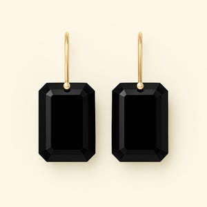 Puede incluir: Un par de pendientes colgantes con piedras negras rectangulares facetadas suspendidas de finos ganchos de metal en tono dorado pulido. Las piedras tienen una superficie lisa y reflectante con bordes biselados, creando un aspecto limpio y simétrico.
