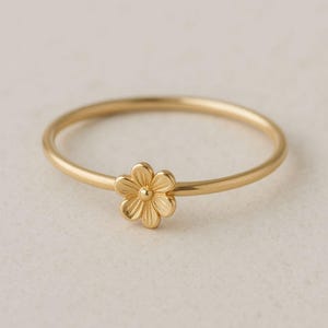 Puede incluir: Un delicado anillo dorado con un pequeño diseño floral detallado. El anillo tiene una banda delgada y un acabado pulido, mostrando una estética minimalista y elegante. La flor está centrada en el anillo, agregando un toque de encanto inspirado en la naturaleza.