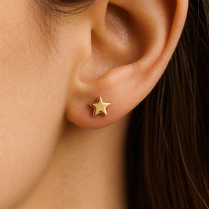 May include: A close-up of a gold star-shaped stud earring on an ear. The earring is a solid, five-pointed star with a polished finish. The skin around the ear is smooth and light-toned.