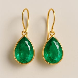 Pode incluir: Um par de brincos pendurados dourados com pedras preciosas verdes esmeralda facetadas em forma de gota. Os brincos têm um design simples com fecho de gancho. As pedras são engastadas em uma moldura dourada.