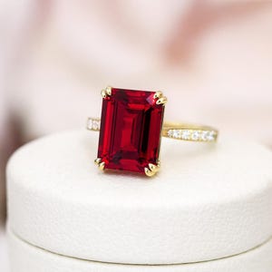 Könnte beinhalten: Ein goldener Ring mit einem großen, rechteckigen, tiefroten Edelstein. Das Goldband ist mit kleinen, klaren Akzentsteinen besetzt. Der Ring wird auf einer weißen Oberfläche mit einem weichen, verschwommenen Hintergrund präsentiert.