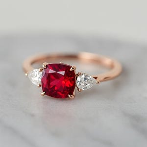 May include: Rose gold ring with a large, square-cut, vivid red gemstone at the centre, accompanied by two pear-shaped, clear gemstones. The ring's band is a delicate, polished rose gold.