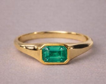 Op de afbeelding: Een gouden ring met een enkele smaragdgeslepen groene edelsteen in een gladde zetting. De ringband is van gepolijst goud met een taps toelopend ontwerp dat naar de centrale rechthoekige steen leidt voor een strak en klassiek sieraad.