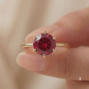 May include: A gold ring featuring a round, faceted deep red gemstone set in a six-prong mounting. The band is smooth and polished, showcasing a classic solitaire design that highlights the vibrant color and clarity of the central stone.