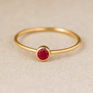 May include: A gold ring featuring a bright red gemstone. The ring has a slender, polished band, offering a minimalist aesthetic. The gemstone is set in a bezel, highlighting its colour. The ring's design is elegant and simple.