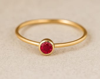 Pode incluir: Um anel de ouro com uma gema vermelha vibrante no centro. A banda é fina e polida, criando um design minimalista e elegante. A gema é engastada em uma moldura, realçando sua cor e brilho.