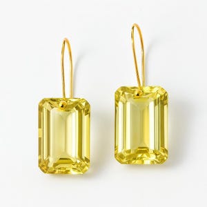 May include: A pair of gold-toned drop earrings featuring large, rectangular, faceted yellow gemstones. The earrings have a simple, elegant design with a hook closure. The gemstones are a vibrant, translucent yellow color.