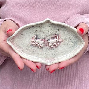 Può includere: Un piatto in ceramica allungato a forma di mandorla con una finitura grigio chiaro maculata e un bordo appuntito. Il centro del piatto presenta due figure di api scolpite in rilievo con sottili accenti rosso rosato sulla superficie.